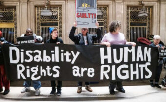 Disabled protestors hold up a dramatic white letter on black banner that says Disability Rights are Human Rights